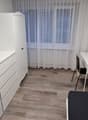 Spacious bedroom with white furniture, including a dresser, desk, and chair. The room has hardwood flooring and large windows with sheer curtains, providing natural light. A small refrigerator and a washing machine are also visible in the room.