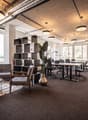 Modern office room with open layout, carpeted floor, gray chairs, tables, glass windows, pendant lighting, and greenery