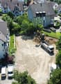 Aerial view of a residential area with a construction site in the center. The site has an excavator and a dump truck, indicating ongoing construction work. The surrounding buildings appear to be multi-story residential apartments with tiled roofs. There a