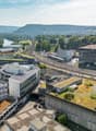 Aerial view of a city with a mix of modern and older buildings, surrounded by hills and greenery. The image shows a train station, parking areas, and green roofs on some of the buildings.