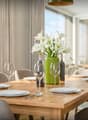 Dining room with wooden table, chairs, white plates, wine glasses, green vase with flowers, wine bottles, cutlery, and windows