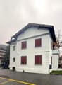 White two-story house, red shutters, beige awning, raised ground floor, entrance to the right