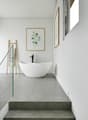 Bathroom with white bathtub, minimalist interior design, grey tiled flooring, glass railing, small window.