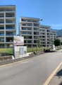 two multistory apartment buildings, white exterior, glass balconies, sidewalk, chain link fence, grassy field, mountain