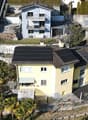 Two-story yellow house with solar panels on the roof, surrounded by other buildings, in a neighborhood with trees.