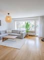 Living room with gray sofa, wooden coffee table, white rug, wooden floor, white walls, large window, potted plant, ceiling lamp, picture frame, and a treadmill.