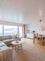 large windows, gray sofa, wooden coffee table, wooden dining table, wooden floor, white walls, ceiling lamp