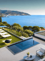 Modern villa with swimming pool, sea view, outdoor lounge area, green lawn, white lounge chairs, black railings, white walls, green plants, round table, potted plant, black lamp