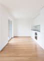 Empty room with wooden floor, white walls, large sliding glass doors, open kitchen, sink, oven.