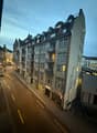 Urban apartment building, facade made of brick, glass windows, multiple floors, balconies, storefronts on the ground floor, traffic lanes, some parked vehicles, and street lamps on the sides.