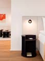 white bed with a blanket and pillows, dark night stand, lit lamp, white walls, picture on the wall, chair and table in the other room