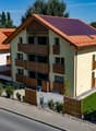 Two-storey house, red roof, yellow exterior, solar panels, wooden balconies, gated entrance, green lawn, road with pedestrian crossing