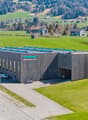 large industrial building with large solar panels on the roof and an entrance with a canopy, cars parked next to the building, and another large building with solar panels at the side