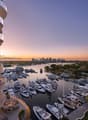 High-rise building with glass balconies, overlooking a marina with many boats and yachts, city skyline in the distance