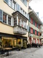 Cobblestone street, buildings with balconies, shops, people walking, outdoor seating, potted plants
