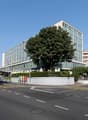 Modern office building, glass facade, multiple floors, surrounded by greenery, street with cars and pedestrians