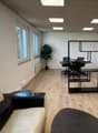 Empty office with wooden floor, white walls, large windows, a couch, a desk, and a potted plant.