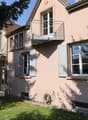 Two story house, pink walls, white shutters, balcony, large tree, garden, patio with table, ground-level access to basement