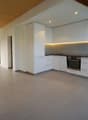Modern, open-concept kitchen with white cabinets, built-in appliances including a dishwasher, and gray tile flooring. The kitchen is integrated into a larger living space with a vaulted ceiling and exposed wooden beams.