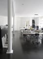 Large open office space with multiple desks, computers, and office equipment. The room has high ceilings, white walls, and dark hardwood floors. There is a dishwasher visible in the corner of the room.
