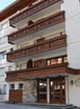 White and brown facade, 3 floors, large balconies with wooden railings, multiple windows, double doors at the entrance