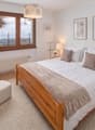 Bedroom with wooden bed, white bedding, wooden nightstands, white chair, white ottoman, white rug, white walls, large window with curtains, radiator, lamps, framed pictures, flowers