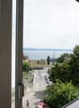 View from the window of a street with cars parked, a person walking, and a building with yellow shutters. In the distance, there is a body of water and a mountain range.