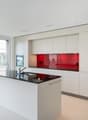Modern, open-concept kitchen with white cabinets, red glass backsplash, black countertop, and built-in appliances including a dishwasher. Large windows provide natural light and access to a balcony or terrace.