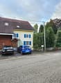 Traditional village house, brown wooden doors, blue shutters, cars parked in front, small garden