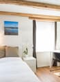 Cozy bedroom with exposed wooden beams, white walls, large window, bed with beige bedding, nightstand, and a desk with a chair.