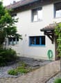 white exterior with brown roof, multiple windows with blue frames, front brick walkway, garden with trees and bushes, hose