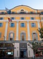 Yellow building with multiple windows, flags, entrance, and shops on the ground floor
