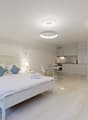 Bed with white sheets and blue pillows, bedside tables with lamps, a blue chair, a kitchen with white cupboards, open floor design, two lamps and a table