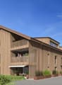 3-story wooden building with a slanted roof, balconies, and large windows. The building has a modern, eco-friendly design with a natural wood exterior. There are people visible on the balconies and a paved walkway in front of the building.