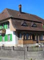 2 story house, brown roof, green shutters, wooden balcony, chimney, concrete wall, wooden fence