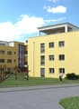 3 story building, yellow paint, white roof, balconies, large windows, play area, sidewalk, lawn, two people walking