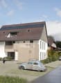 Modern house with solar panels, white exterior, brown roof, driveway with cars, person on bicycle, trees, and greenery.