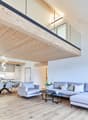 Spacious open-plan living area with wooden ceiling beams, large windows, modern kitchen with white cabinets, dining table with chairs, and a comfortable gray sofa set with coffee tables.