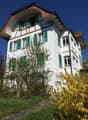 Two-story house with a traditional Swiss-style architecture, featuring a red-tiled roof, white walls, and green shutters. The house has a balcony or terrace on the upper level. The surrounding garden is lush with trees and shrubs, including a yellow flowe