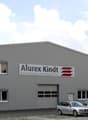 Large gray industrial building with the Alurex Kindt logo, multiple windows, and a parking lot in front of the building