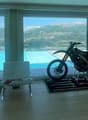 Large open-concept living room with floor-to-ceiling windows offering a panoramic view of the mountains and a swimming pool outside. The room features a modern design with a dirt bike displayed on a platform in the center.