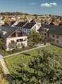 The image shows a scenic residential area in a hilly, forested region. The buildings are a mix of multi-story apartment buildings and smaller, single-family homes with red tile roofs. The buildings have balconies and are surrounded by lush greenery, inclu