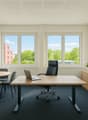 Spacious office with large windows providing a view of the surrounding greenery, a wooden desk with a laptop, chairs, and shelves with plants and office supplies.