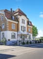 2 story house, brown roof, brick walls, windows with shutters, front garden, metal fence, stairs to the entrance, sign on the wall