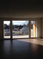 Spacious living room with large windows overlooking a balcony or terrace, dark tile flooring, and a view of the surrounding buildings and landscape outside.