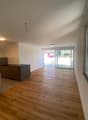 Open-plan living area with hardwood flooring, white walls, large windows providing natural light, and a sliding glass door leading to a balcony or terrace.