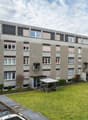 4 story apartment building, grey exterior, multiple windows, bicycles parked in front, small garden area