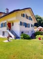 2 story house, yellow, white windows, blue shutters, red door, stairs to entrance, paved driveway, garden, flower pots, fence, small gazebo