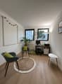 small room with wooden floor, white walls, two chairs, round rug, desk with computer, radiator, plant, clock, picture frame