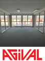 Large open office space with floor-to-ceiling windows providing a view of the surrounding buildings. The room has a gray carpet floor and a desk chair visible in the corner.